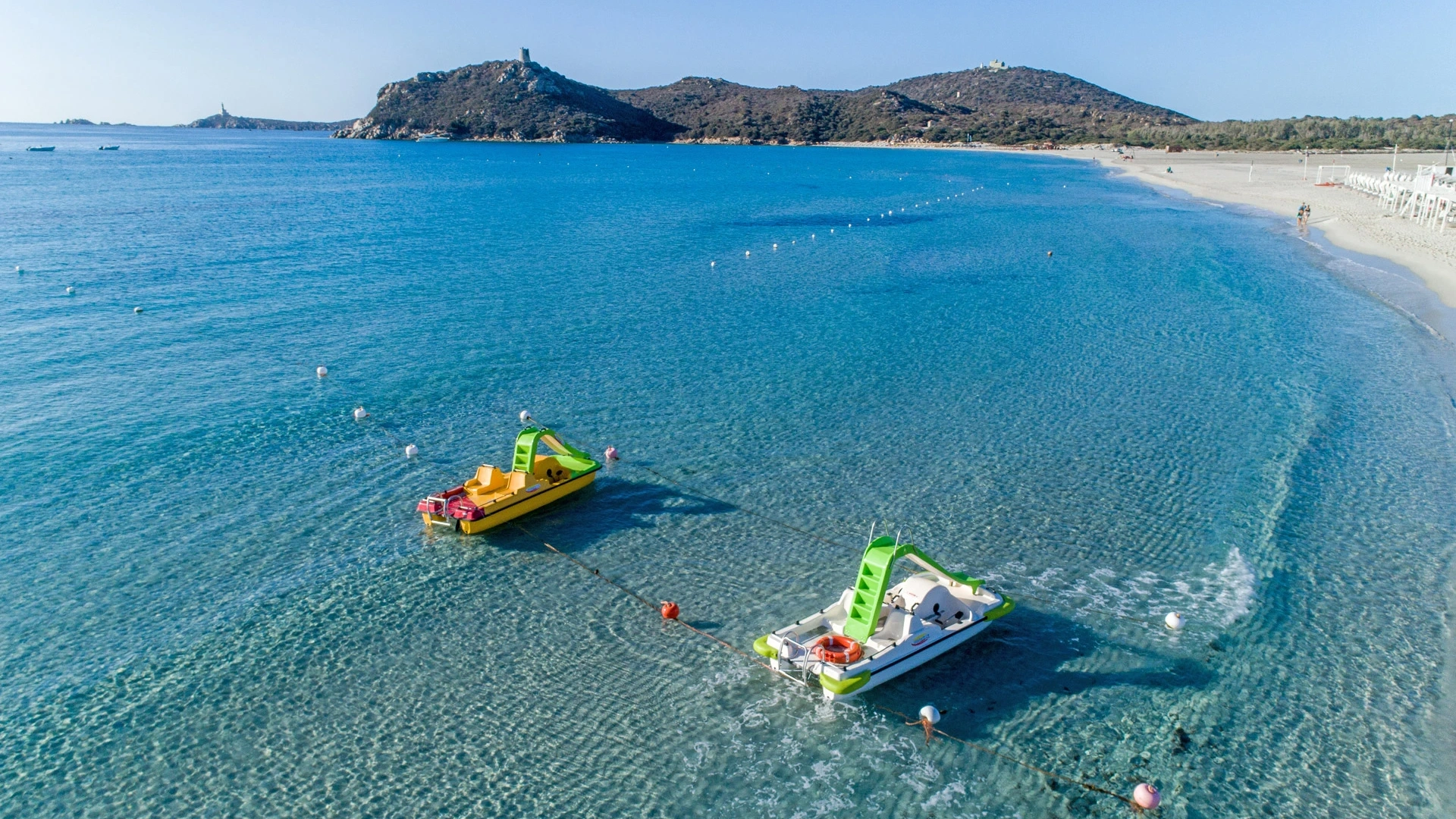 Mare cristallino con pedalò colorati, spiaggia e colline in lontananza.