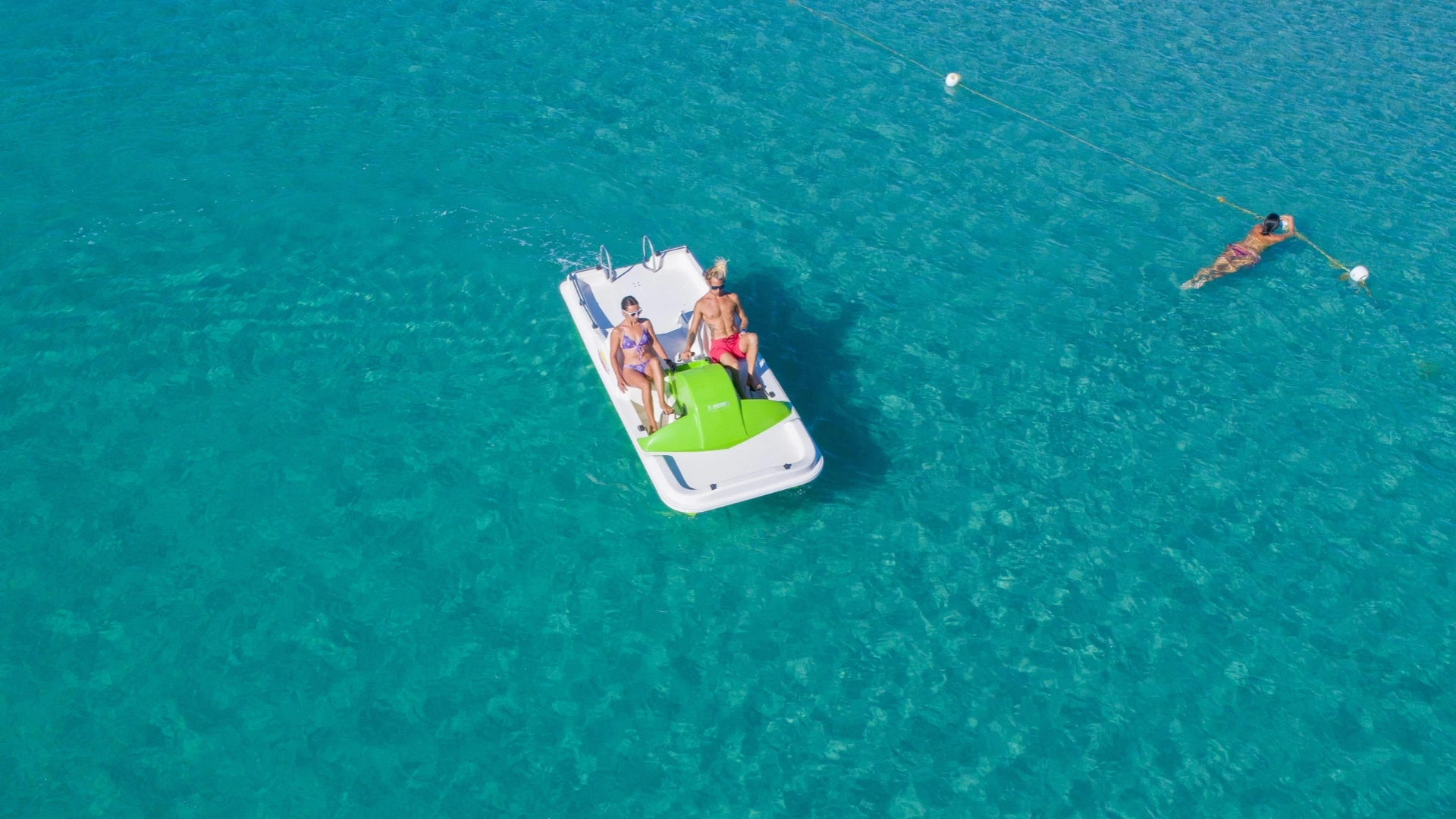 Due persone su un pedalò verde in mare, una nuota vicino alla corda.