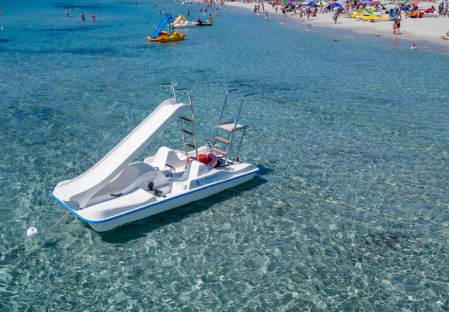 Pedalò con scivolo in mare cristallino, spiaggia affollata sullo sfondo.